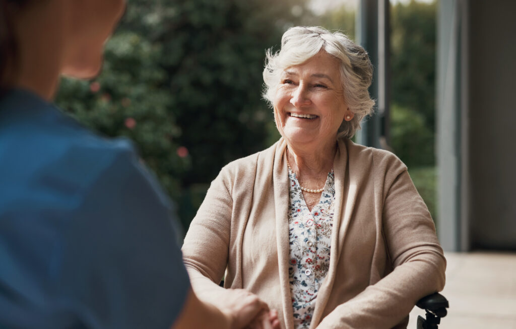 Holding hands, nurse and happy senior woman in a wheelchair with help or support for wellness. Elderly patient person with a disability and caregiver talking about healthcare, smile or medical care